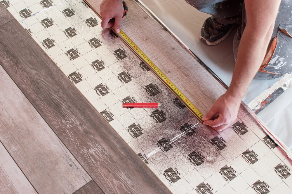 Personne installant un revêtement de sol, agenouillée sur un plancher en bois, utilisant un mètre ruban jaune pour mesurer une lame de parquet