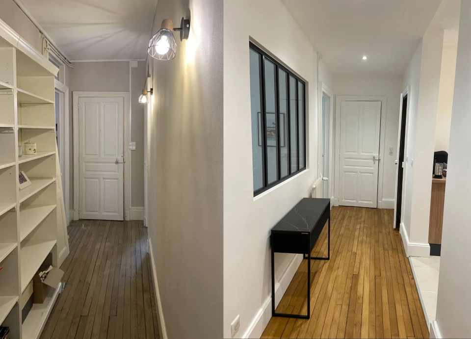 Un artisan en tenue de travail professionnelle (chantier) pose ou finit l'installation d'une porte intérieure blanche dans un cadre de rénovation intérieure. La scène montre l'artisan en train de régler ou fixer le dormant de la porte, avec en arrière-plan une pièce en cours de rénovation (murs peints, parquet visible). Qualité photo réaliste et professionnelle, éclairage naturel, perspective qui met en avant l'artisan et son savoir-faire.