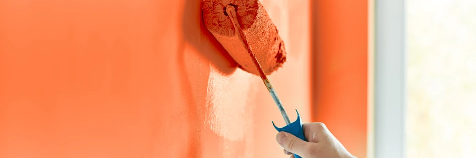 Peintre professionnel en tenue blanche et casquette appliquant de la peinture orange sur un mur intérieur à l'aide d'un rouleau à poils longs